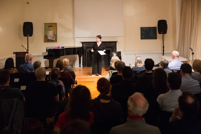 Anabella Lenzu in black stands in front of seated audience. There is a piano and fireplace in the back ground